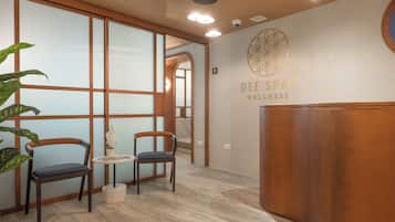 Un área de recepción con un escritorio de madera, dos sillas, una pequeña mesa con un florero y una pared con el logotipo 'DEE SPAS WELLNESS'.