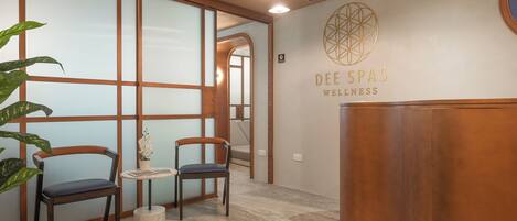Un área de recepción con un escritorio de madera, dos sillas, una pequeña mesa con un florero y una pared con el logotipo 'DEE SPAS WELLNESS'.