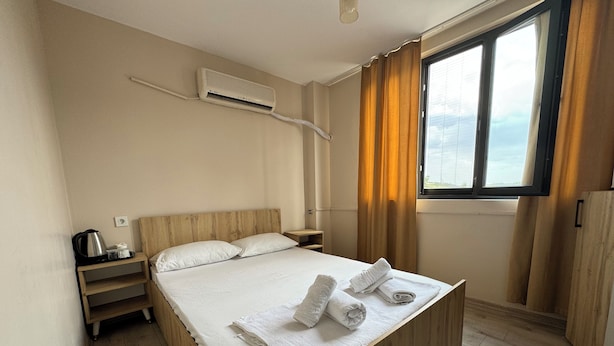 A neatly made bed with white linens and towels, a wooden headboard, a bedside table with a kettle, and a window with curtains. -  Avio Hotel (Diyarbakir)