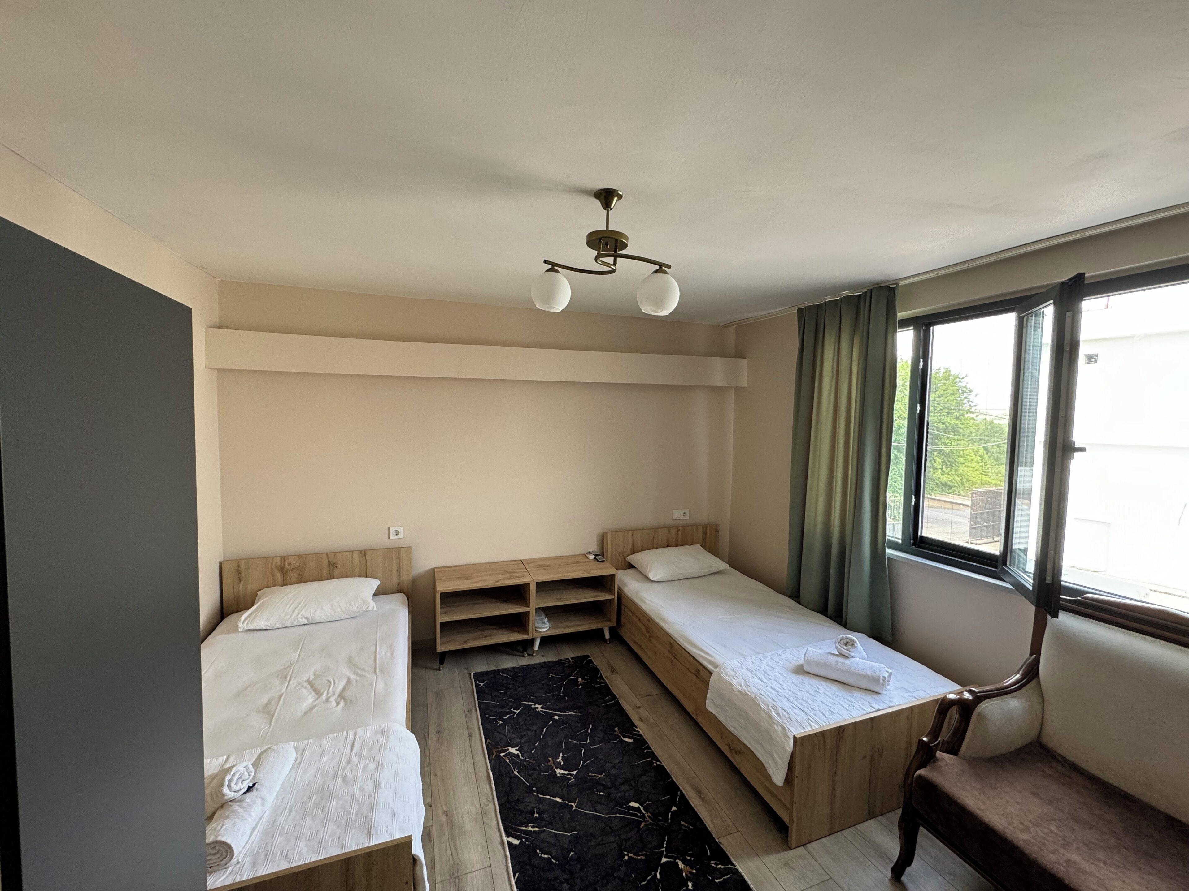 A small, well-lit room with two single beds, a wooden bench, a small table, and a ceiling light fixture.