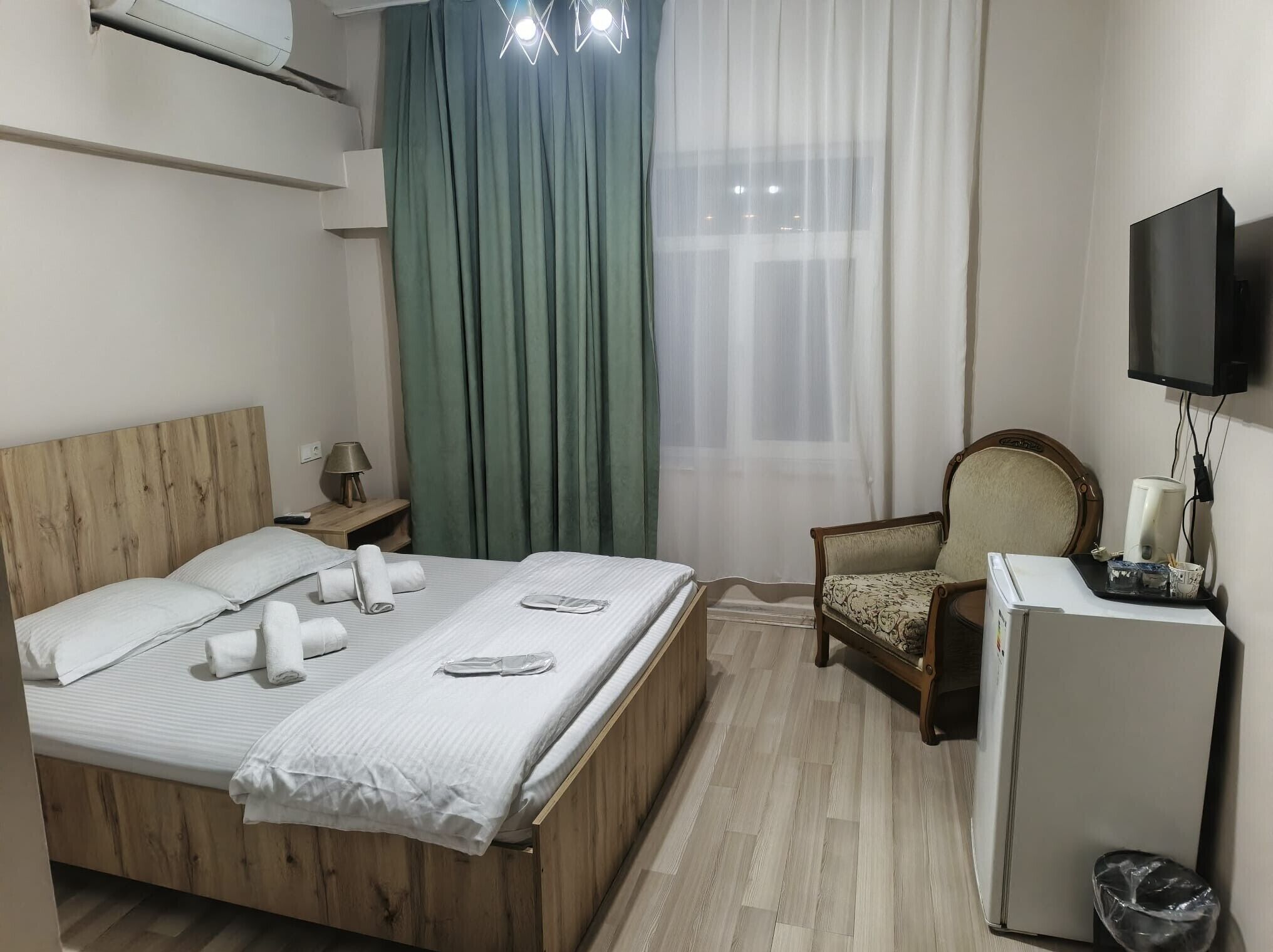 A hotel room with a wooden bed, a small desk with a lamp, a chair, a television, and a refrigerator.