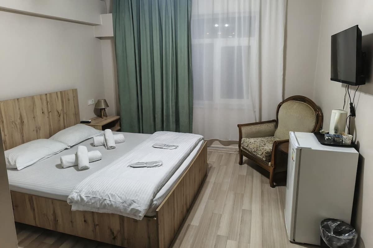 A hotel room with a wooden bed, a small desk with a lamp, a chair, a television, and a refrigerator.