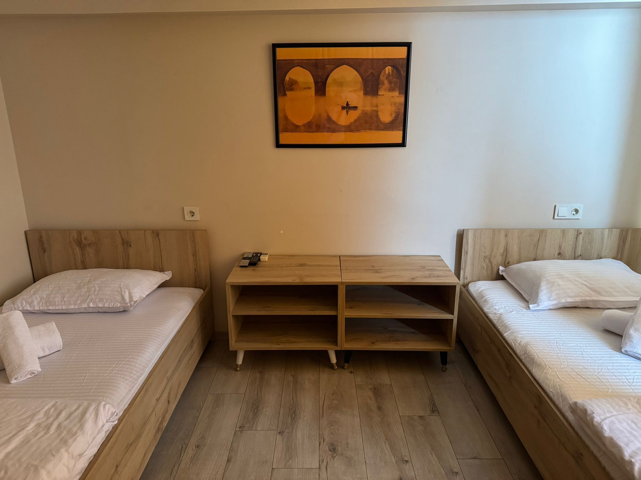 A room with two single beds, a wooden nightstand with a remote control, and a framed picture on the wall.