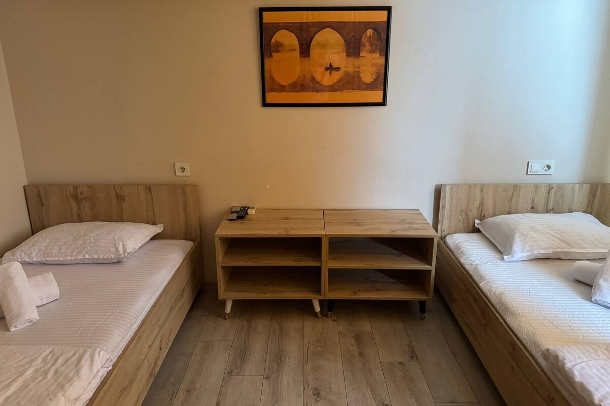 A room with two single beds, a wooden nightstand with a remote control, and a framed picture on the wall.