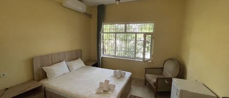 A bedroom with a wooden bed, a small bedside table, a chair, a desk, and a window with a view of greenery.