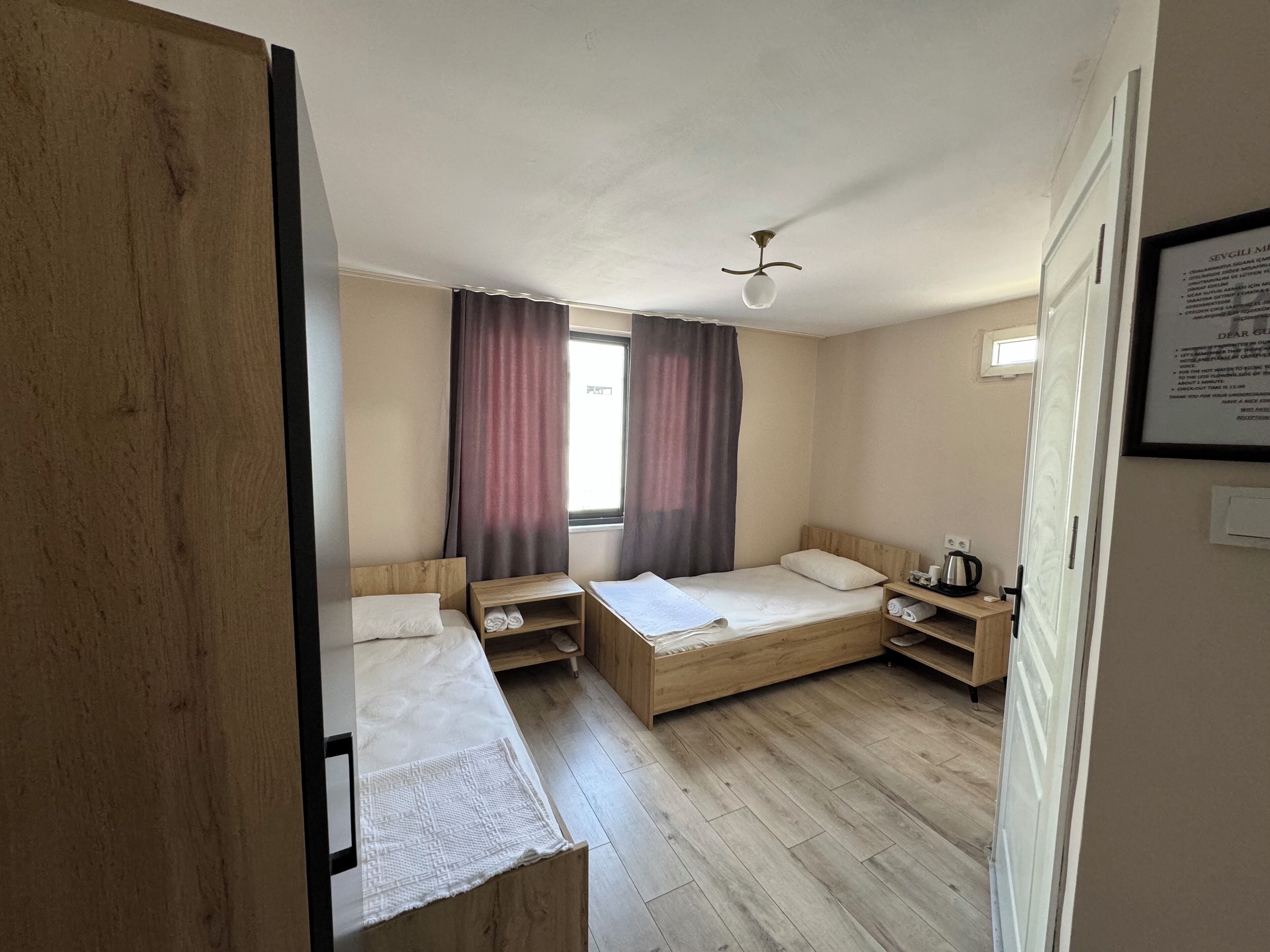 Habitación de hotel con dos camas individuales, cabecera de madera, una mesita pequeña, una mesita de noche, una ventana con cortinas y un ventilador de techo.