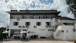 A two-story building with a sign reading "HOME'S HOTEL". There are air conditioning units on the exterior, a covered outdoor seating area with tables and chairs, and a tiled patio. -  Avio Hotel (Diyarbakir)
