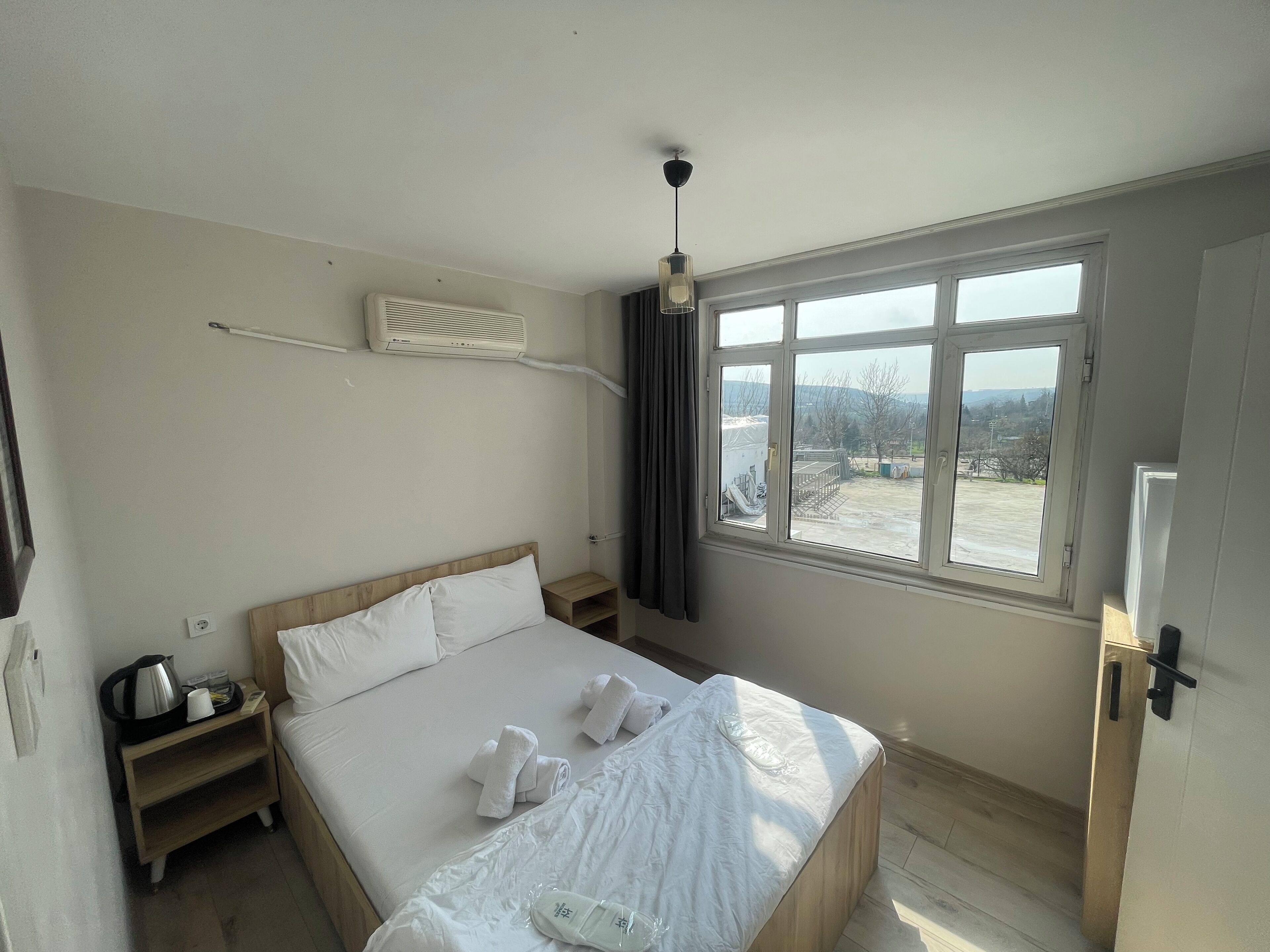 A hotel room with a bed, a bedside table, a heater, and a window with a view of the outside.