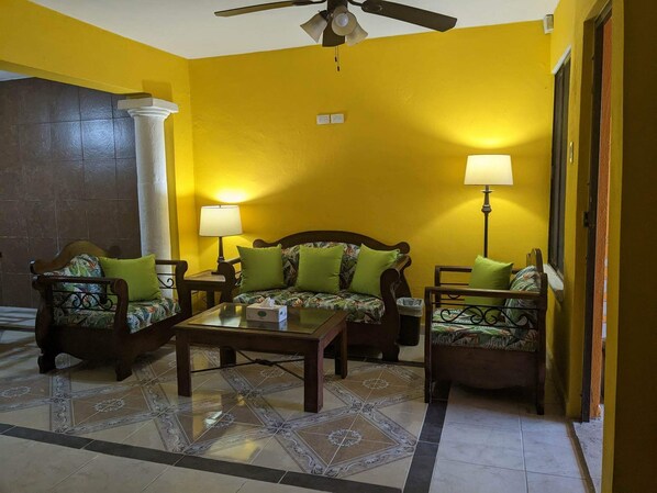 A living room with yellow walls, a ceiling fan, and two lamps. There is a sofa and two chairs with green cushions. - Beautiful Casa De Rose (Progreso)