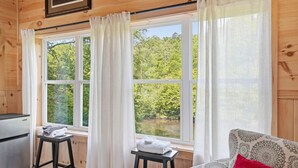 A cabin with wooden walls, a kitchenette, a sofa with a patterned cushion, and a view of a forest through large windows. - The Riverside Cabin! (Blue Ridge)