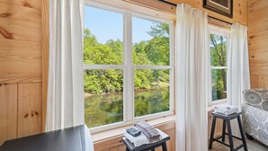 A cabin with a large window offering a view of a lake and forest. There is a small table with towels and a chair. - The Riverside Cabin! (Blue Ridge)