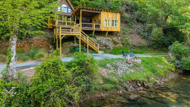 A wooden cabin with a staircase leading to a deck, situated by a river surrounded by greenery. - The Riverside Cabin! (Blue Ridge)