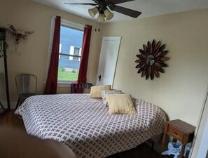 Ein Schlafzimmer mit Bett, Deckenventilator, Fenster mit Vorhängen, einem Sessel und Wanddekoration.