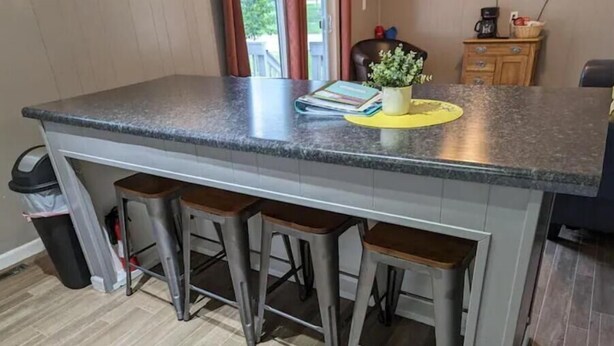 A kitchen with a granite countertop, three bar stools, a yellow placemat, and a potted plant. - Home Away From Home (Biggsville)