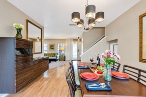 A dining area with a wooden table set for four, a chandelier, and a view of the living room in the background.