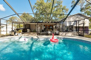 A swimming pool with a glass enclosure, surrounded by a patio area with outdoor furniture and a barbecue setup. - Stunning Home w/ Heated Saltwater Pool & Pond View + 12 Guests + Great Location (Temple Terrace)