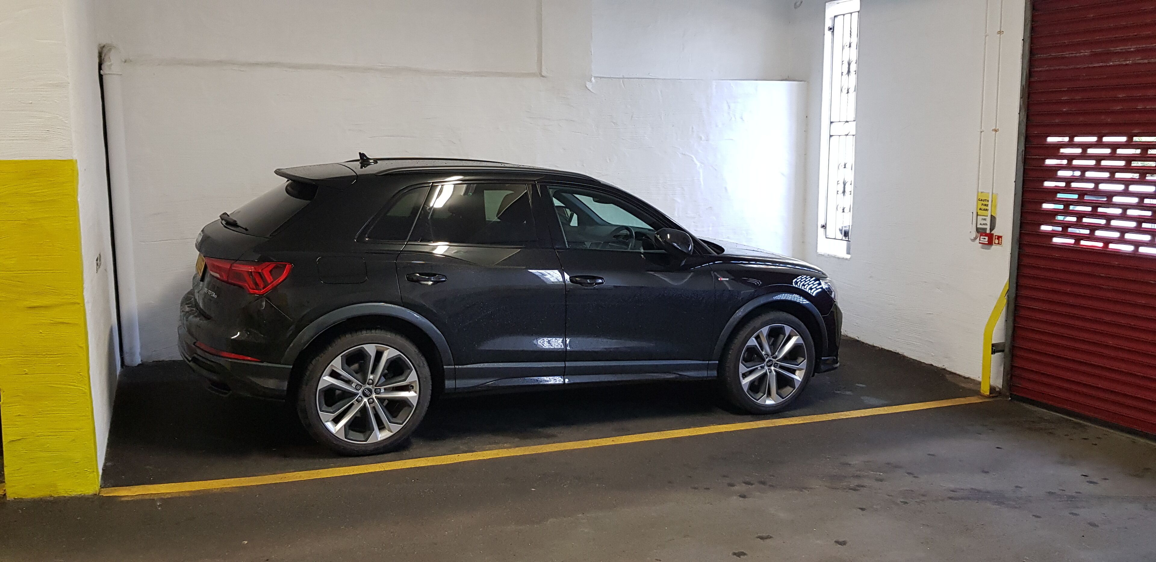 A black SUV parked inside a garage with a yellow line on the floor, a red garage door, and a white wall with a window.