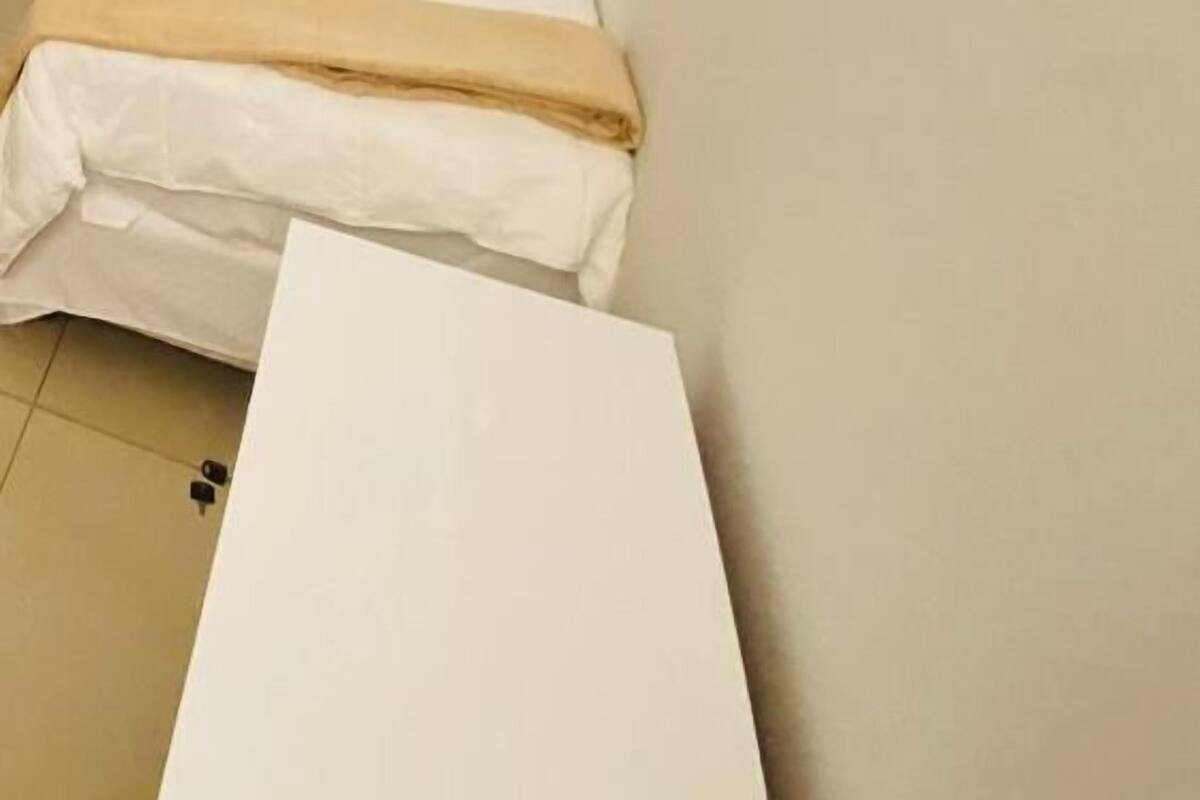 Ein kleines Zimmer mit einem Einzelbett, einem schlichten weißen Bett und einer beigen Decke. Das Zimmer hat ein schlichtes, zweckmäßiges Design, eine schlichte Wand und ein kleines Fenster.