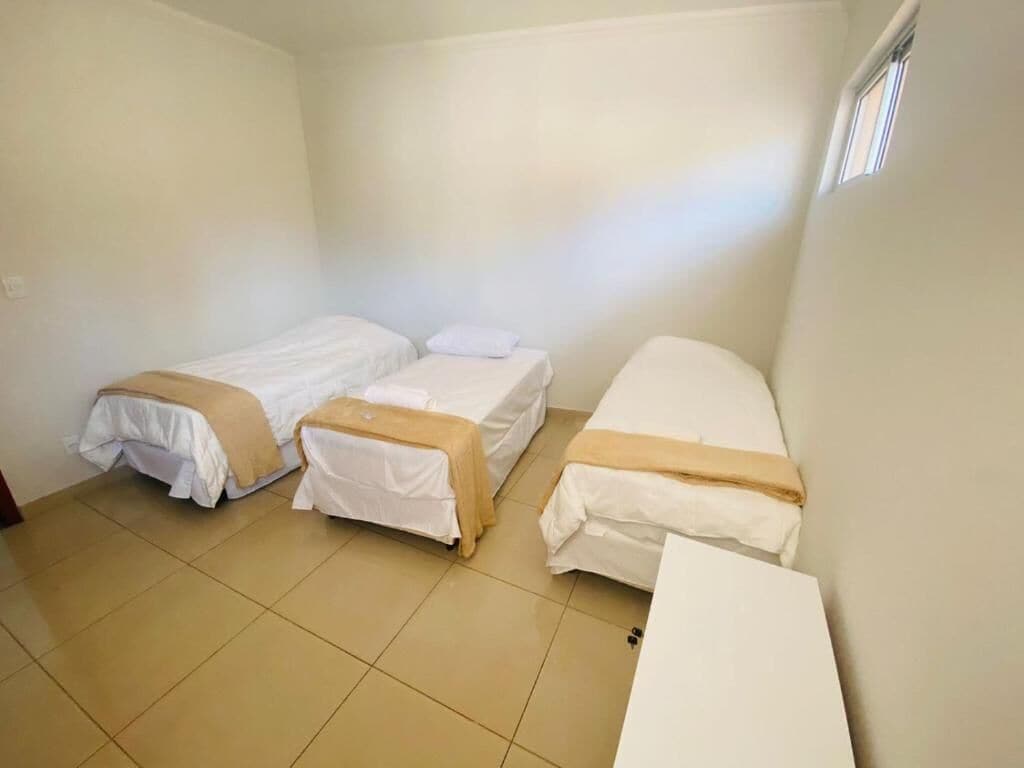 A small, clean room with three single beds, a white desk, and a window providing natural light.