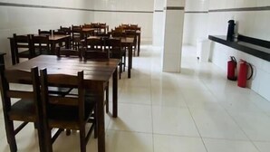 A spacious dining area with multiple wooden tables and chairs, a white tiled floor, and a kitchen area with a black countertop and red fire extinguishers.