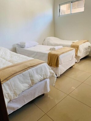 Une chambre avec trois lits simples, chacun doté d’un matelas blanc et d’une couverture marron. La chambre a un sol carrelé et une fenêtre donnant sur l’extérieur.