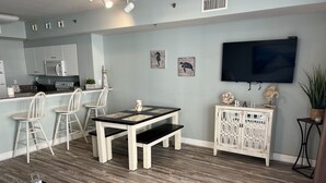 A modern kitchen with white cabinets, a microwave, and a sink. There is a dining area with a table and chairs, and a TV mounted on the wall. - Gorgeous Sunset Condo at One of the Most Beautiful Pool Resorts on the Beach!  (Panama City Beach)