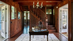 A wooden interior with a staircase, a chandelier, and a table with books and a bicycle.