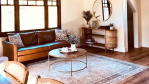 A spacious living room with a high ceiling, a ceiling fan, a large window, a sofa, a coffee table, and a mirror. - Villa Verde - 5 miles to Disney! (Fullerton)
