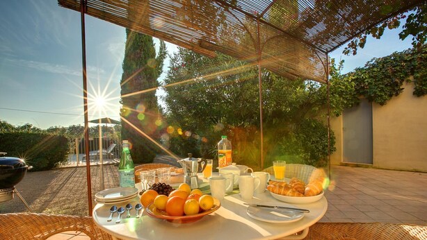 An outdoor breakfast setup with a table, wicker chairs, and a variety of breakfast items including fruits, bread, and beverages.