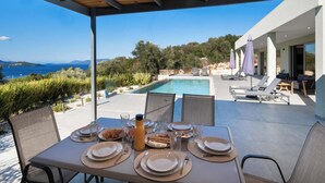 A covered outdoor dining area with a pool and lounge chairs, overlooking a scenic landscape with a view of the sea and hills. - Kristi - Three Bedroom Villa, Sleeps 6 (Spartochori)
