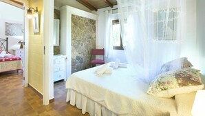 A bedroom with a stone wall, a bed with a canopy, a nightstand, a chair, and a window with curtains.
