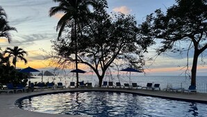 A pool area with lounge chairs and umbrellas, surrounded by trees and a view of the sea at sunset. - BEAUTIFUL VIEWS ON PLAYAS GEMELAS BEACH (PUERTO VALLARTA)