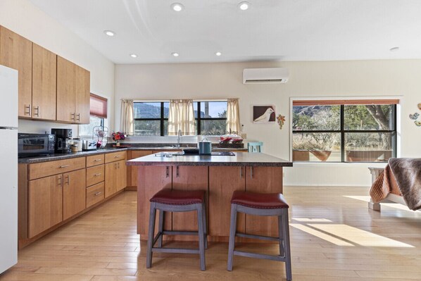 A modern kitchen with wooden cabinets, a central island with stools, a large window with curtains, and a view of the outdoors.