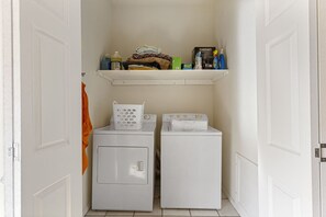 Une buanderie avec deux machines à laver blanches, une étagère contenant divers objets et une porte sur laquelle est accrochée une serviette orange.
