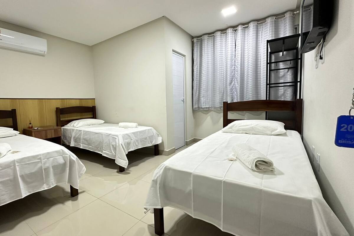 A hotel room with three single beds, a wooden headboard, a white air conditioning unit, a window with curtains, and a sign indicating the room number.