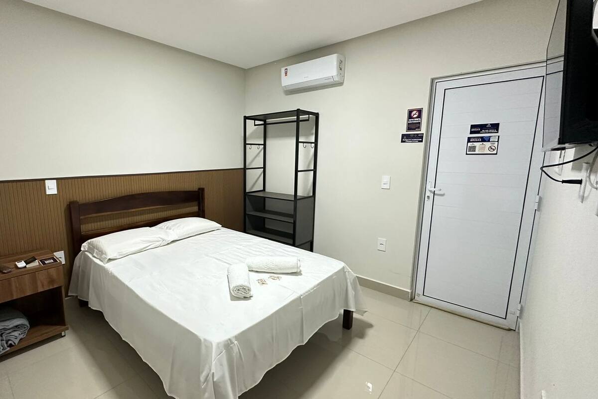 A hotel room with a bed, a wooden headboard, a shelf with toiletries, a wardrobe, and an air conditioning unit.