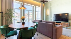 A modern living room with a round wooden table, green chairs, a sofa, a television, and a balcony with a view of buildings.