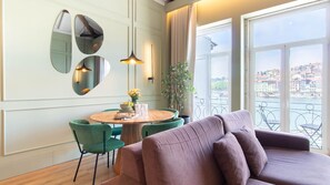 A modern living room with a sofa, a dining table with chairs, a plant, and a view of a cityscape through large windows. - Riverside Oasis by LovelyStay (Vila Nova de Gaia)