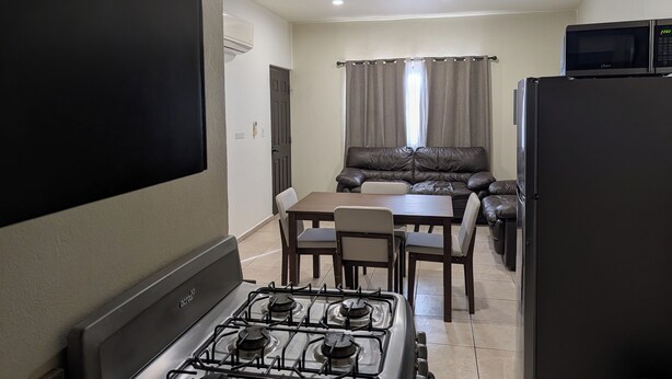 A compact kitchen with a gas stove, a refrigerator, a dining table with chairs, a sofa, and a TV mounted on the wall. - Cómodo Departamento Y Para Cuatro Personas, Cerca del Aeropuerto de Loreto (Loreto)