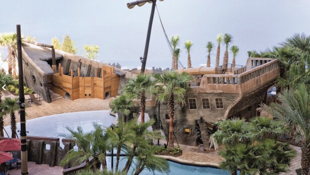 Un área de piscina con una estructura de barco de madera, palmeras y un puente de piedra.