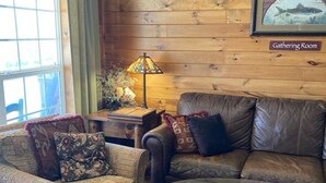 A cozy room with a wooden wall, a mounted deer head, a framed picture of a trout, a couch, a glass coffee table, and a patterned rug.