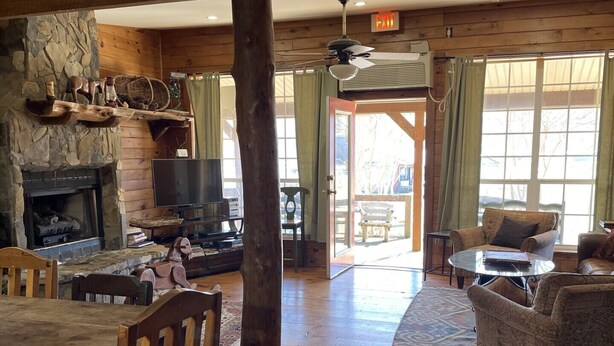 A rustic lodge interior with a stone fireplace, wooden furniture, and a large window providing natural light.