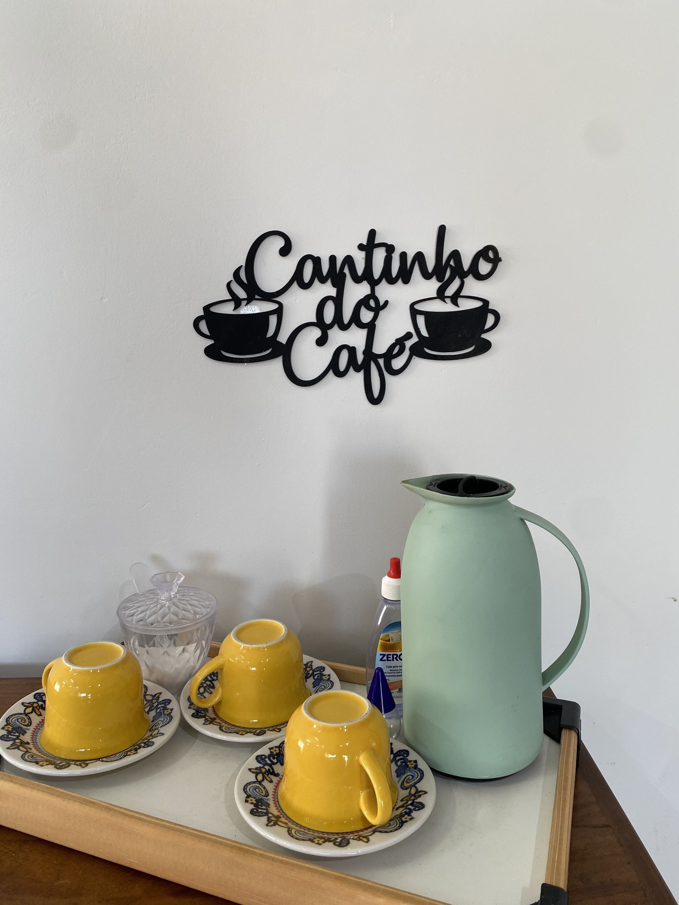 Wanddecoratie met de tekst 'Cantinho do Cafe' en een ontwerp van een koffiekop. Vier gele koffiekopjes staan op een dienblad met schoteltjes, een glazen vaas en een groene kan.