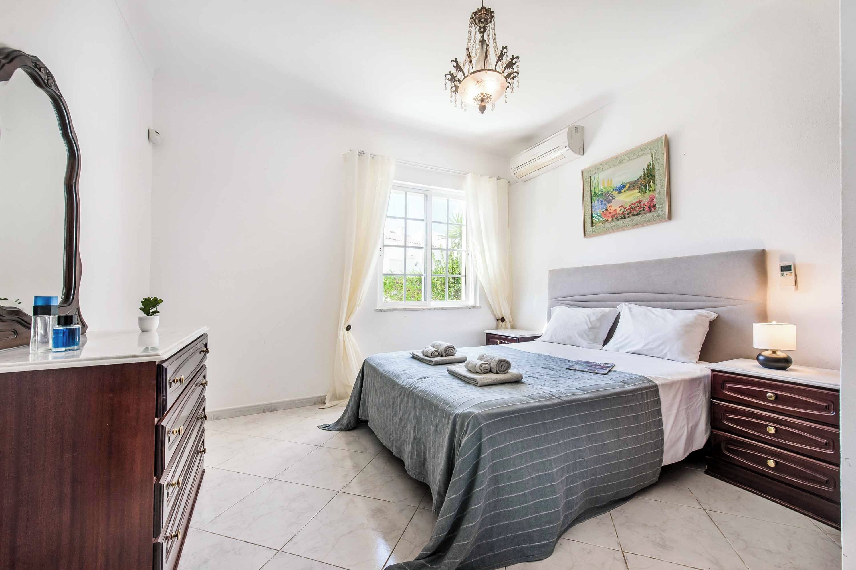 A bedroom with a bed, bedside table, lamp, dresser, and a window with curtains.