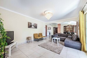 A modern living room with a flat-screen TV, two armchairs, a sofa, a coffee table, and a chandelier.