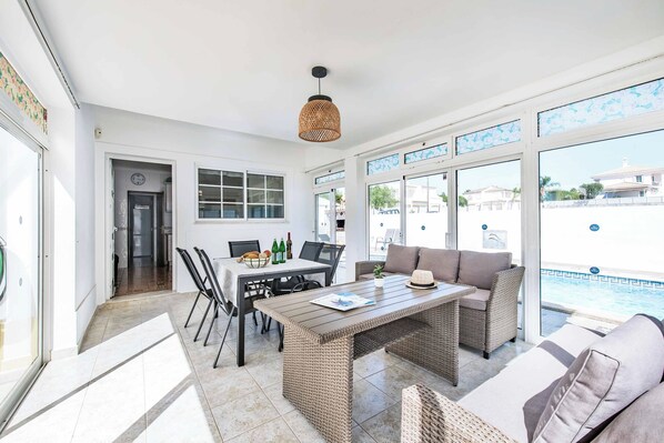 A sunlit room with a dining table, chairs, a wicker sofa, and a view of a swimming pool.