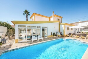 A poolside area with a clear blue pool, white lounge chairs, and a building with large windows and a tiled roof.