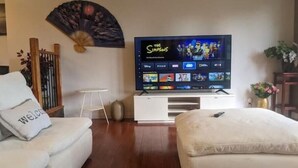A living room with a flat-screen TV displaying 'The Simpsons', a white sofa with a 'Welcome' pillow, a small white side table, and a decorative fan on the wall.