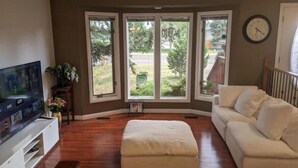 A living room with a large flat-screen TV, a white sofa, a beige ottoman, and a wooden coffee table. There are three large windows with a view of a backyard.