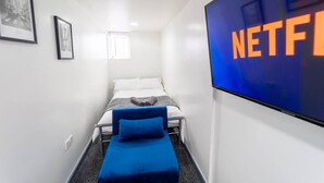 A narrow hotel room with a bed, a blue chair, a TV screen displaying "NETFLIX," and two framed pictures on the wall.